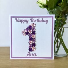 A white square card standing on a shelf next to a vase of flowers. A light purple background and white foreground with purple wording saying Happy Birthday at the top and Ava at the bottom. A large number one is in the middle filled with hand punched flowers of 3 different colours.