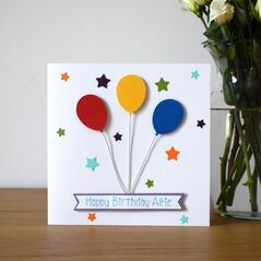 A white square card featuring 1 red, 1 blue and 1 yellow balloon with silver string leading down to a white and lilac banner with the words happy birthday and a child's name. Surrounded by stars of two different sizes in the colours green, turquoise, orange and purple.